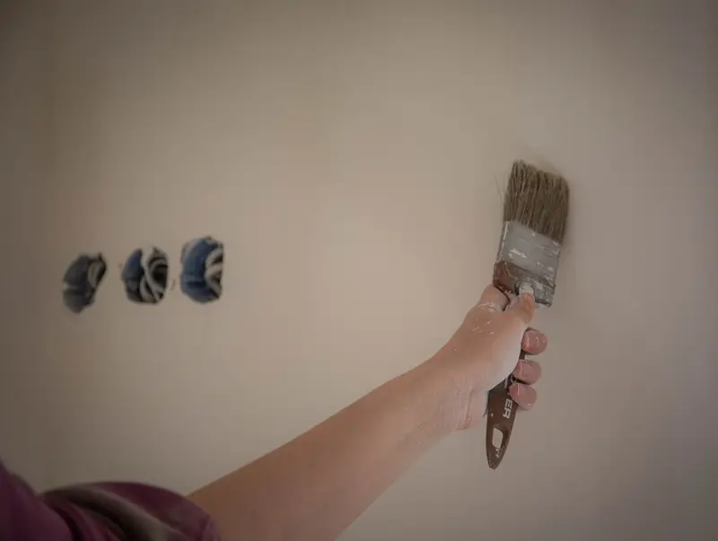 Een hand met een kwast boven een beige muur.