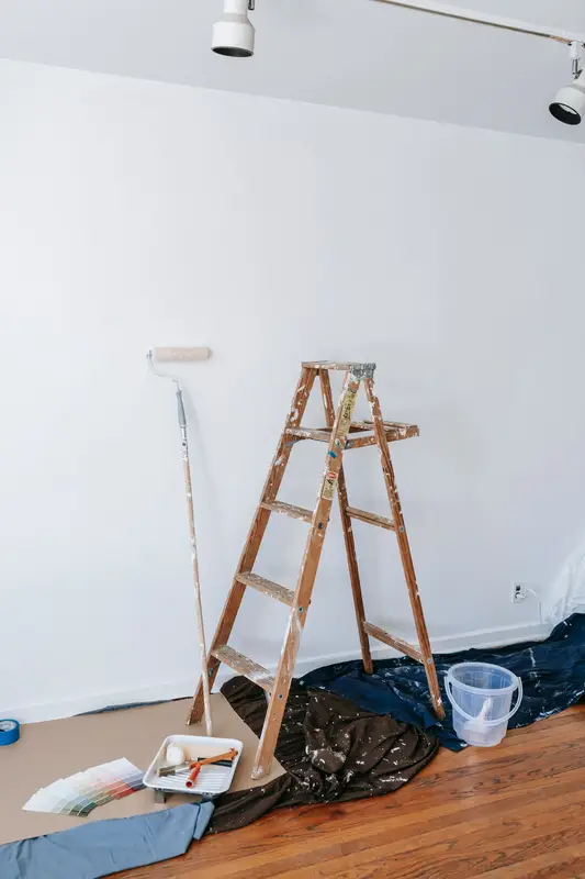 Een schildersladder en roller in een lege kamer met witte muren.