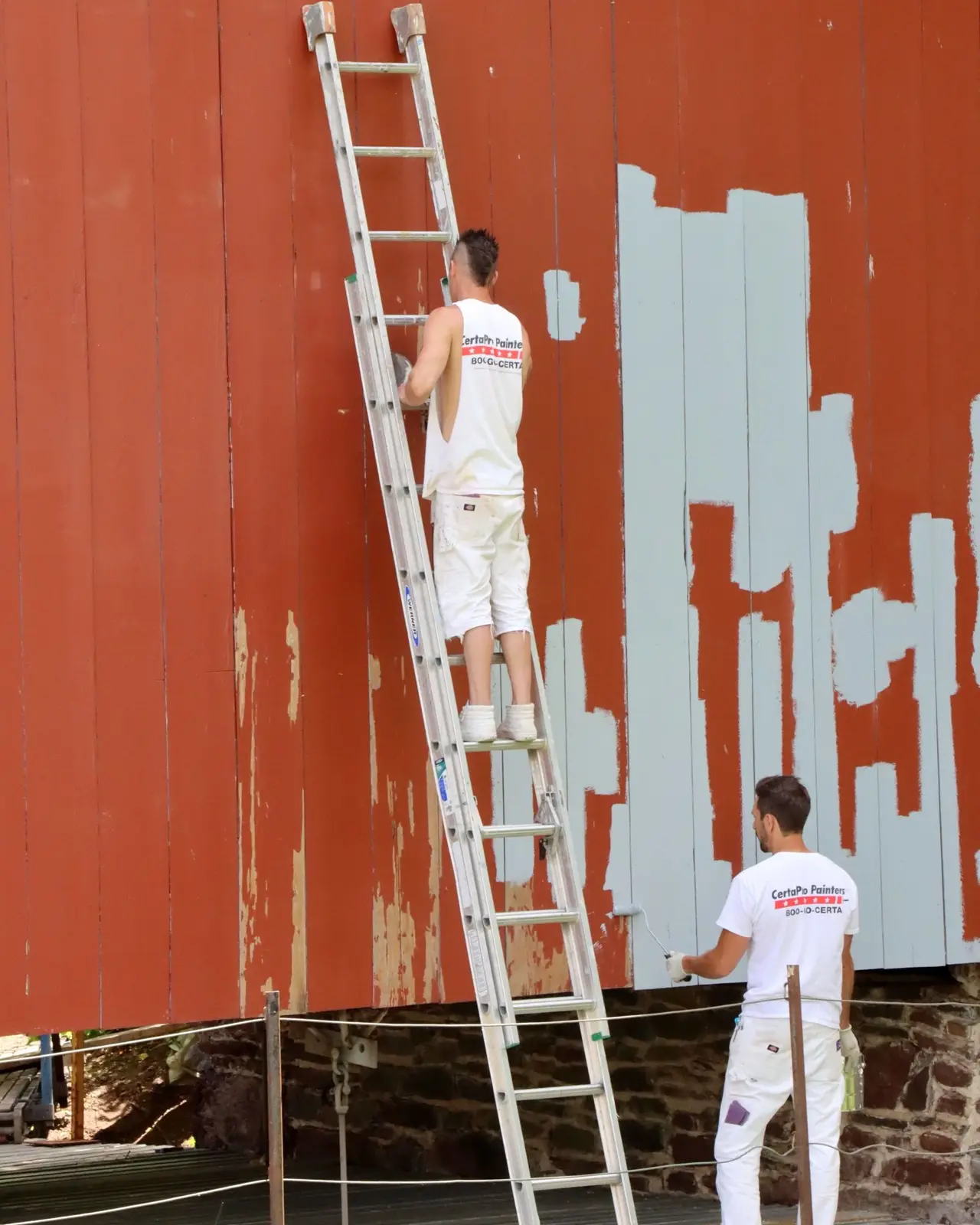 Twee schilders op een ladder zetten een rode houten gevel in grijsblauw.